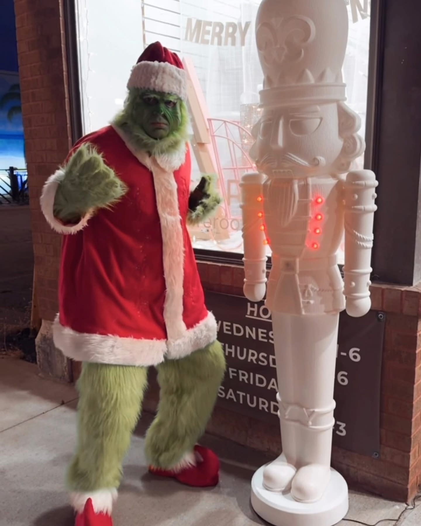 The Grinch in a Santa suit posing aggressively next to a tall, white decorative nutcracker statue inside a store or event space.