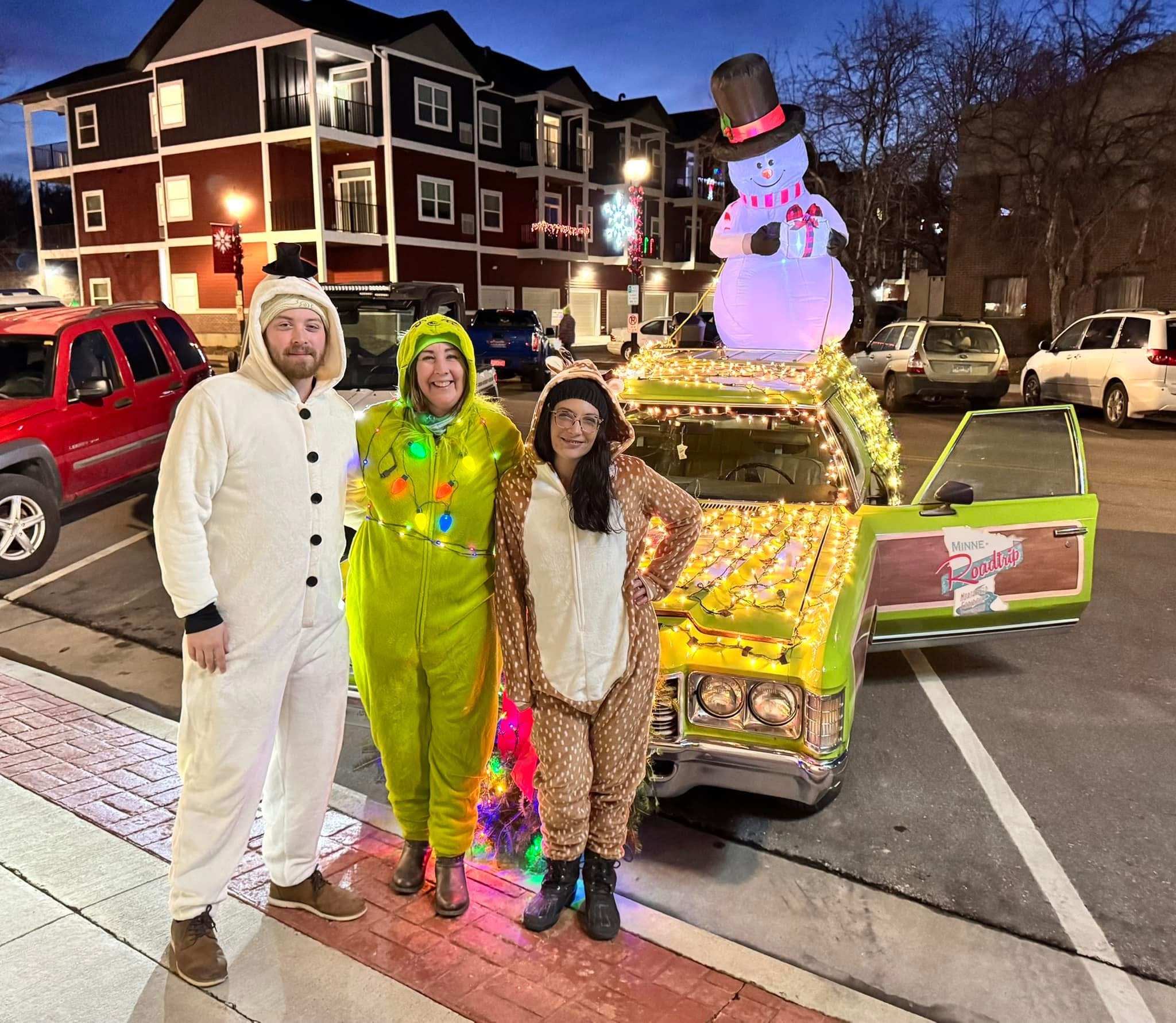 Three people dressed in holiday costumes—a snowman, the Grinch, and a reindeer—posing next to a vintage car heavily decorated with yellow lights and an inflatable snowman.