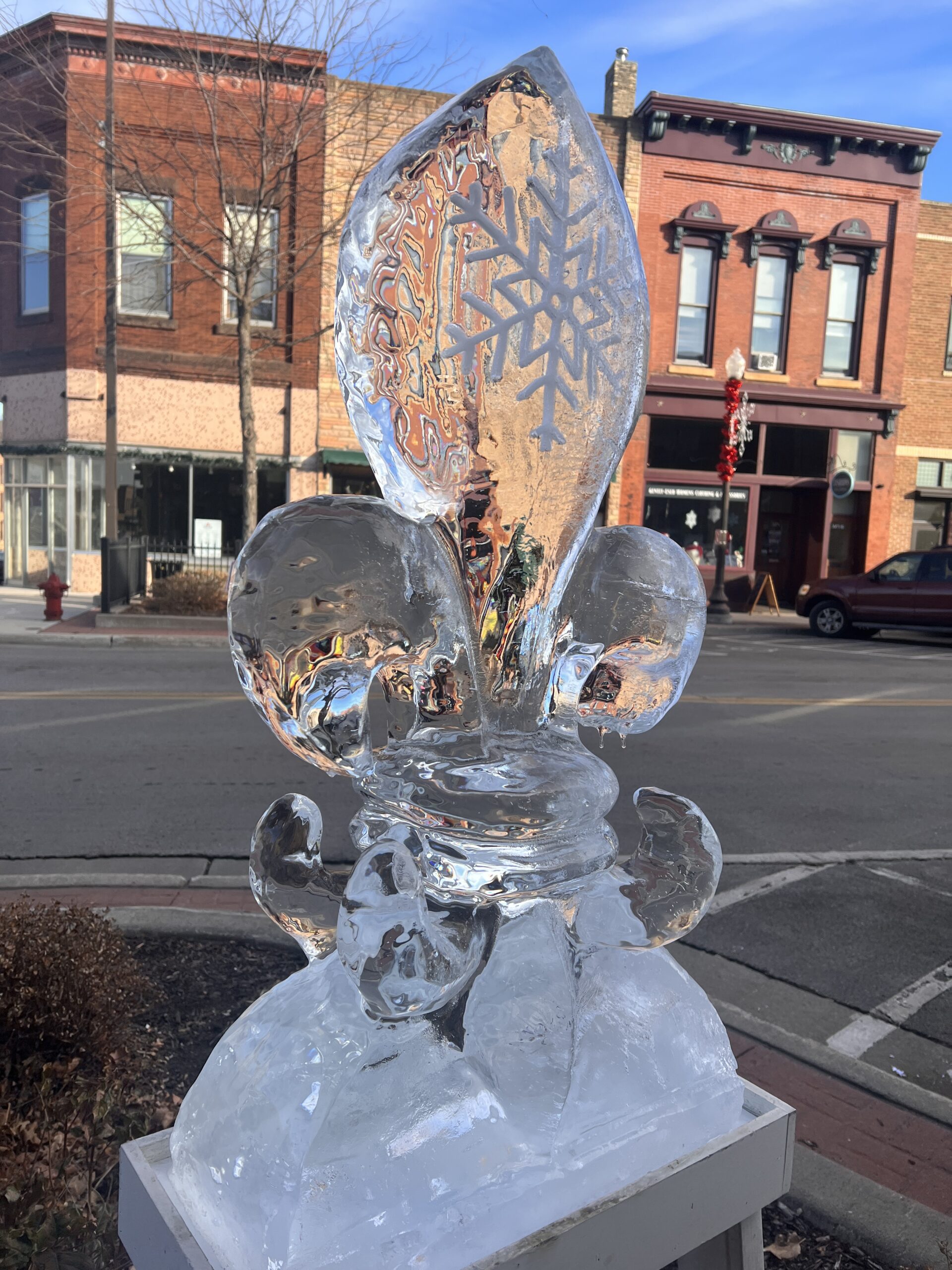 Ice carving of a fleur-de-lis with a snowflake design carved into the center, sitting on a pedestal in downtown Faribault.