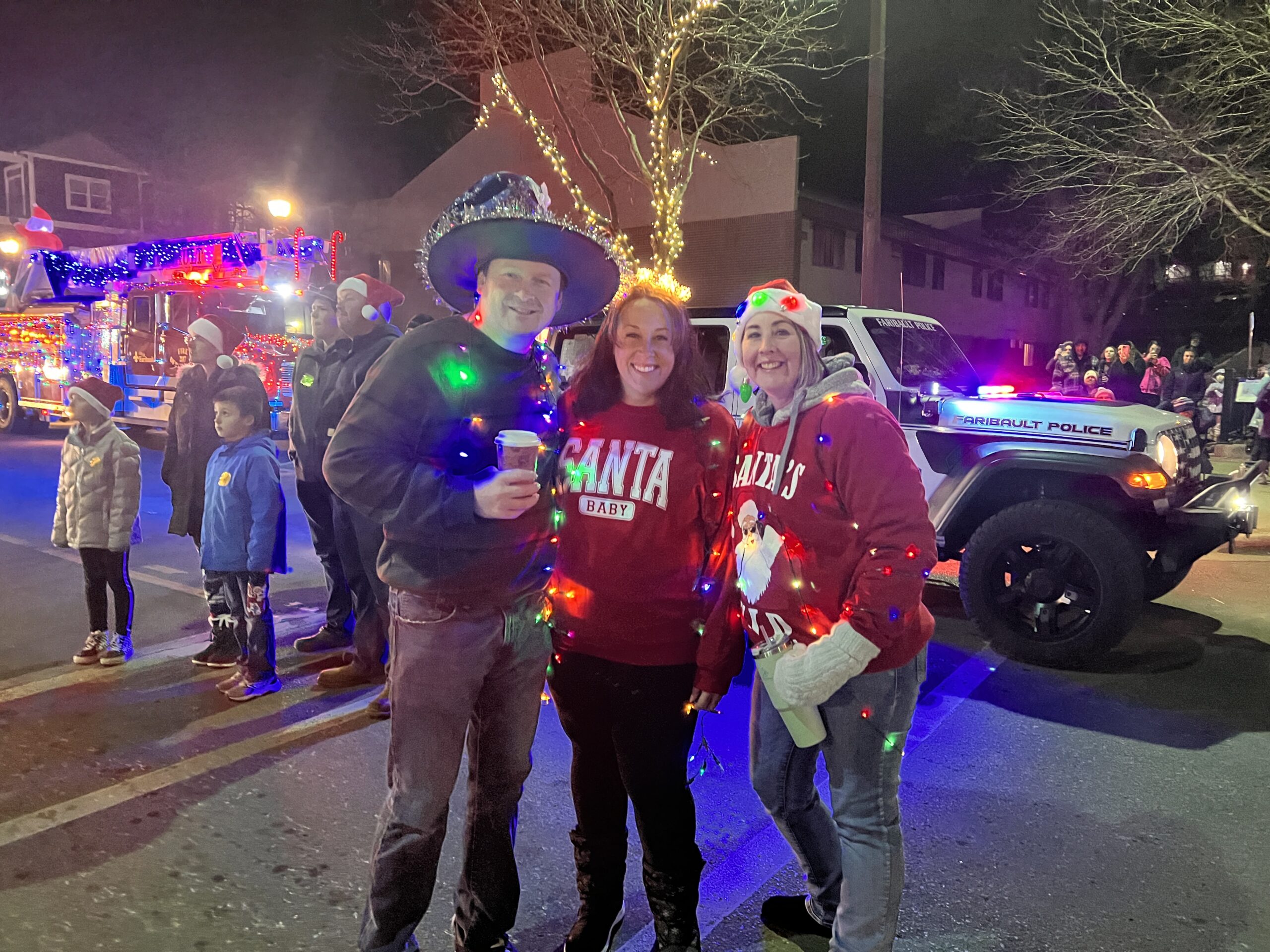 Three friends smiling and posing with festive lights wrapped around them, wearing "Santa Baby" and "Ho Ho Ho" sweaters in front of a Faribault Police vehicle and parade floats.