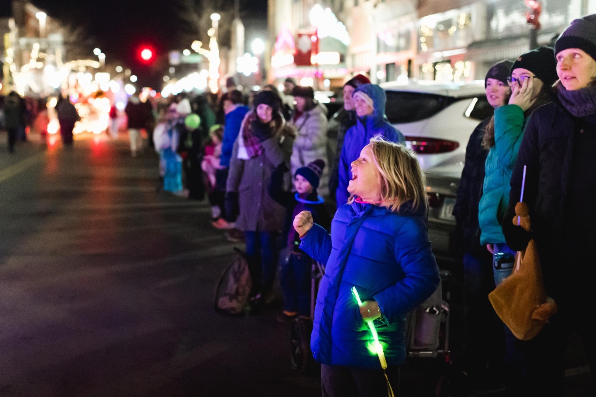 A young girl in a blue winter coat smiling with excitement while holding a glowing green light stick, watching the night parade alongside a group of spectators.